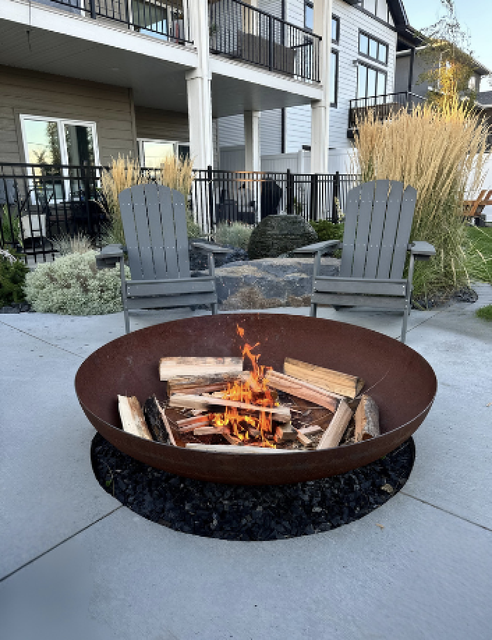 Steel fire pit bowl with burning logs on black rock mulch, Adirondack chairs and basalt sphere water feature behind